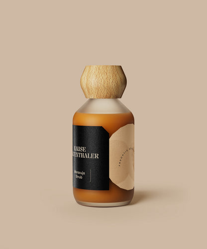 Eine braune Flasche mit Holzkappe und schwarzem Etikett, auf dem MARACUJA SHRUB von Haus Kaltenthaler abgebildet ist, vor einem beigen Hintergrund.
