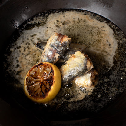 SARDINEN IN AZOREN-BUTTER von José Gourmet brutzeln in einer Pfanne mit einer gerösteten Zitronenhälfte.