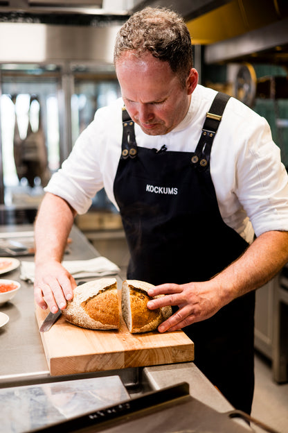 In einer professionellen Küche schneidet der Koch fachmännisch einen runden Laib Brot auf einem robusten MASSIVES KÜCHENBRETT AUS SCHWEDEN von Kockums Jernverk und stellt dabei Präzision und Können unter Beweis.