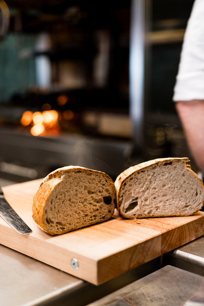Zwei Hälften handwerklich hergestellten Brotes liegen auf einem MASSIVES KÜCHENBRETT AUS SCHWEDEN von Kockums Jernverk, in gemütlicher Küchenatmosphäre liegt ein Messer in der Nähe.