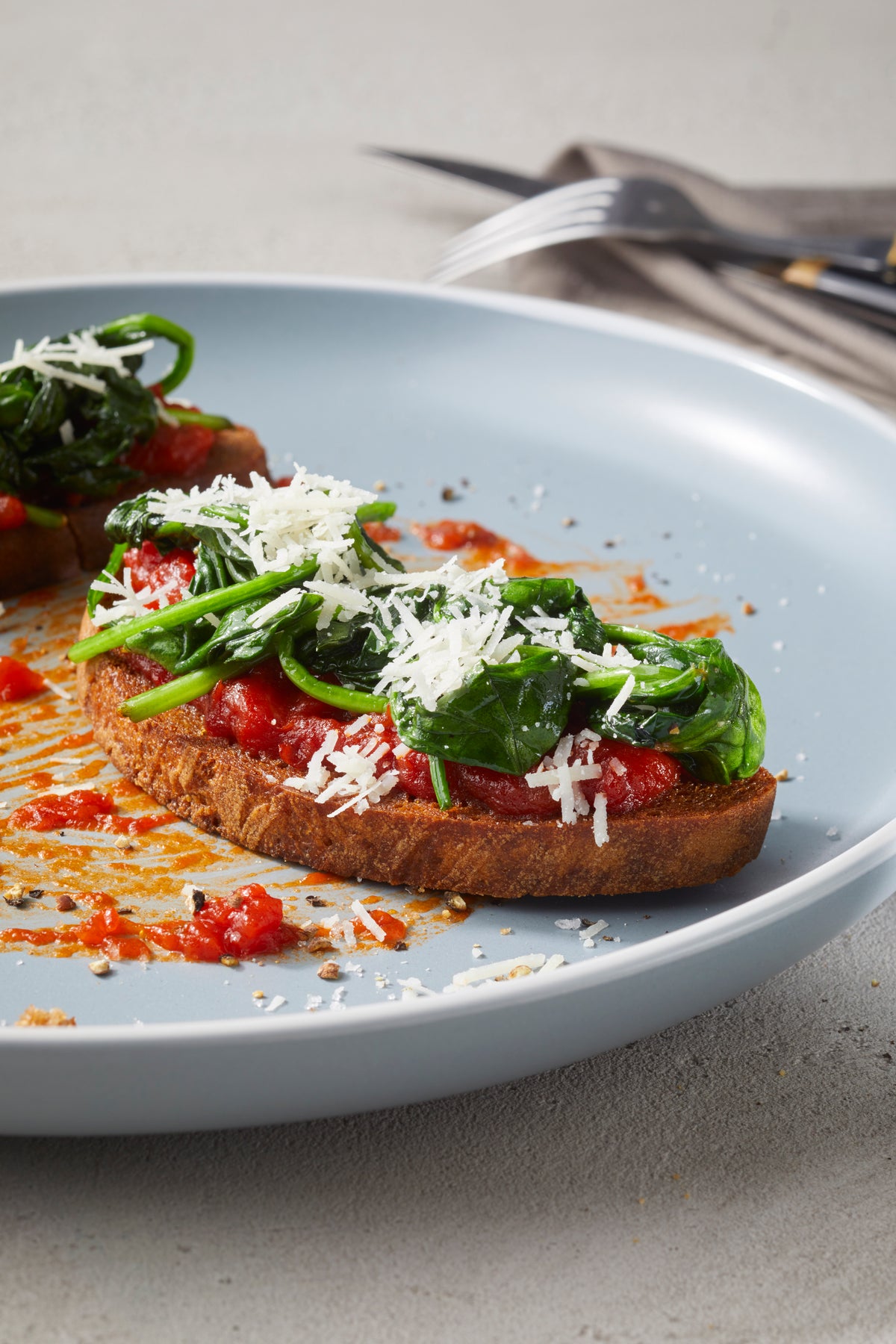 Eine Scheibe Toast mit Rittmeyers TOMATE FRITO IN OLIVENÖL, sautiertem Spinat und geriebenem Käse auf einem hellblauen Teller.