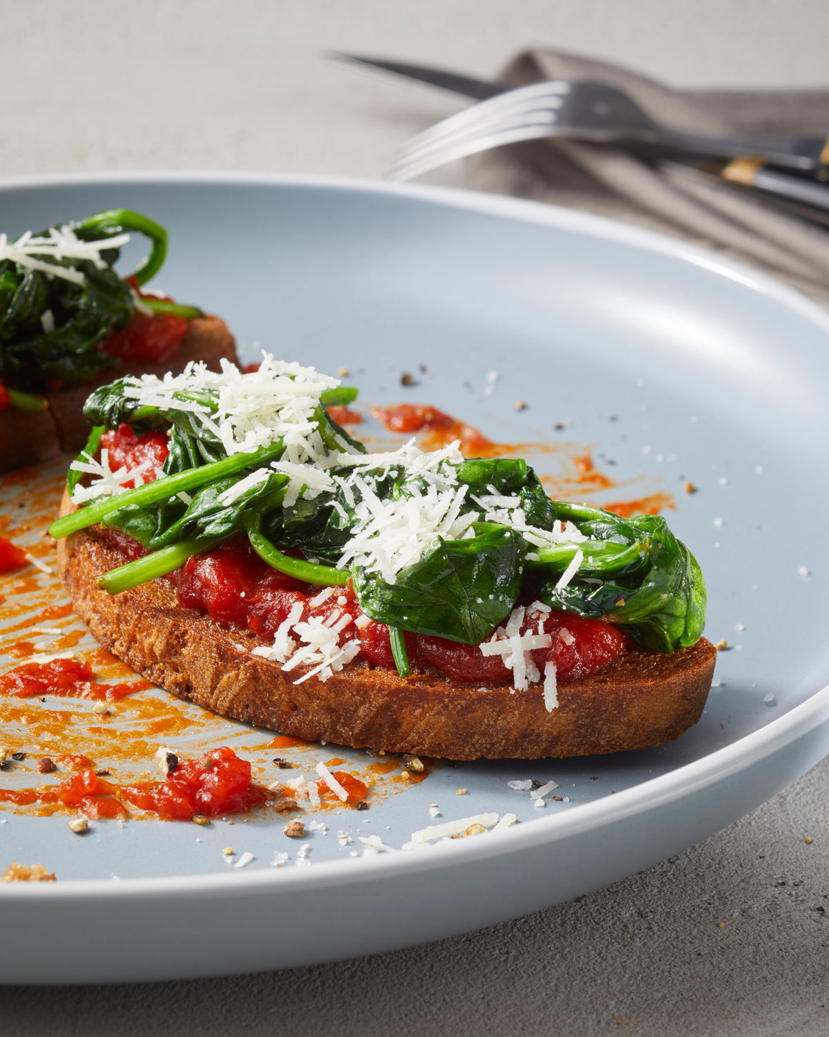Eine Scheibe Toast mit Rittmeyers TOMATE FRITO IN OLIVENÖL, sautiertem Spinat und geriebenem Käse auf einem hellblauen Teller.