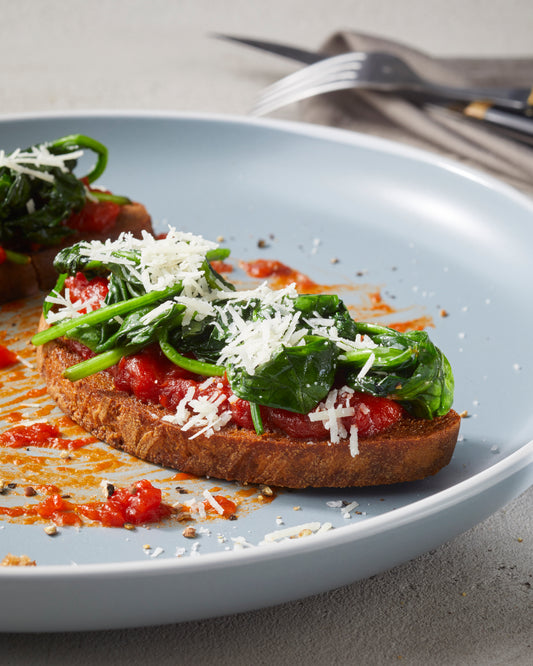 Eine Scheibe Toast mit Rittmeyers TOMATE FRITO IN OLIVENÖL, sautiertem Spinat und geriebenem Käse auf einem hellblauen Teller.
