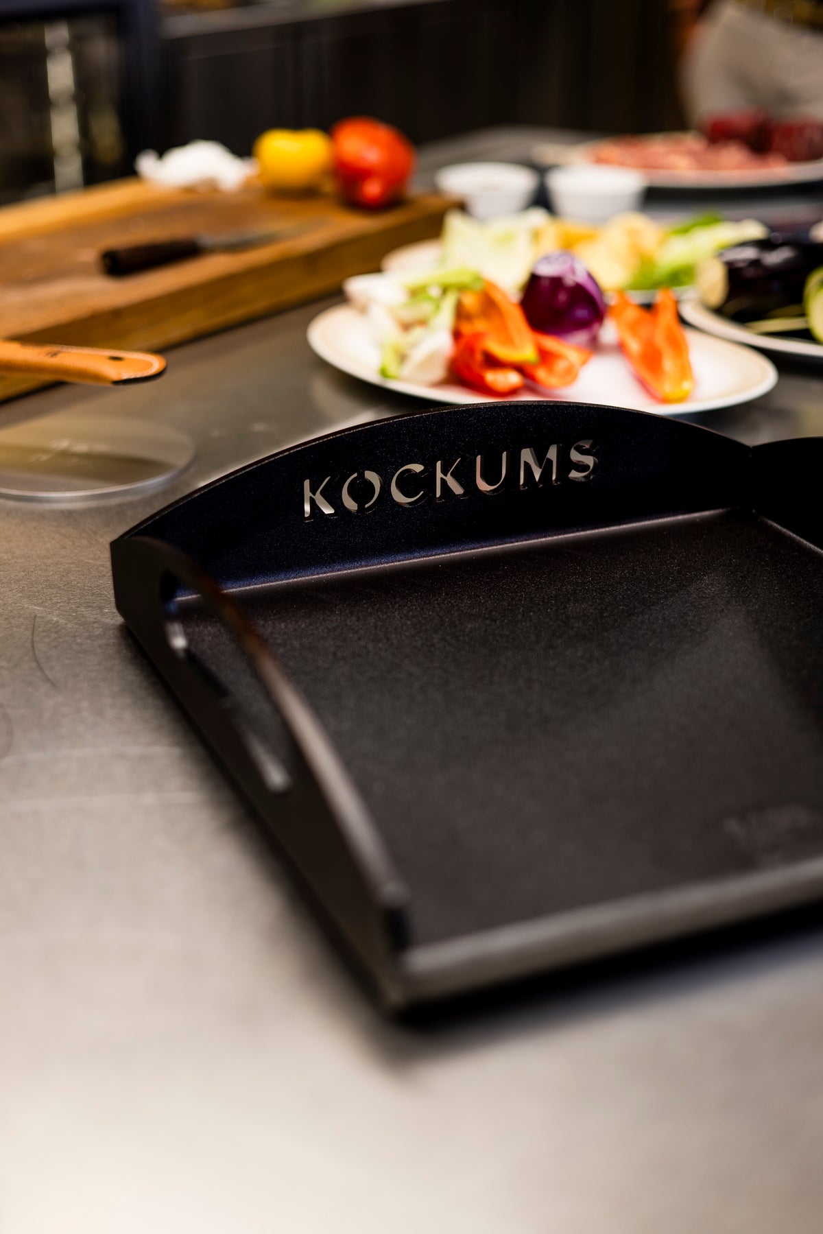 Eine Kockums Jernverk GRIDDLEPLATTE 35 x 35 cm, aus schwedischem Kohlenstoffstahl gefertigt und dezent mit dem Kockums-Logo versehen, steht auf einer Küchentheke mit verschwommenem Gemüse im Hintergrund.