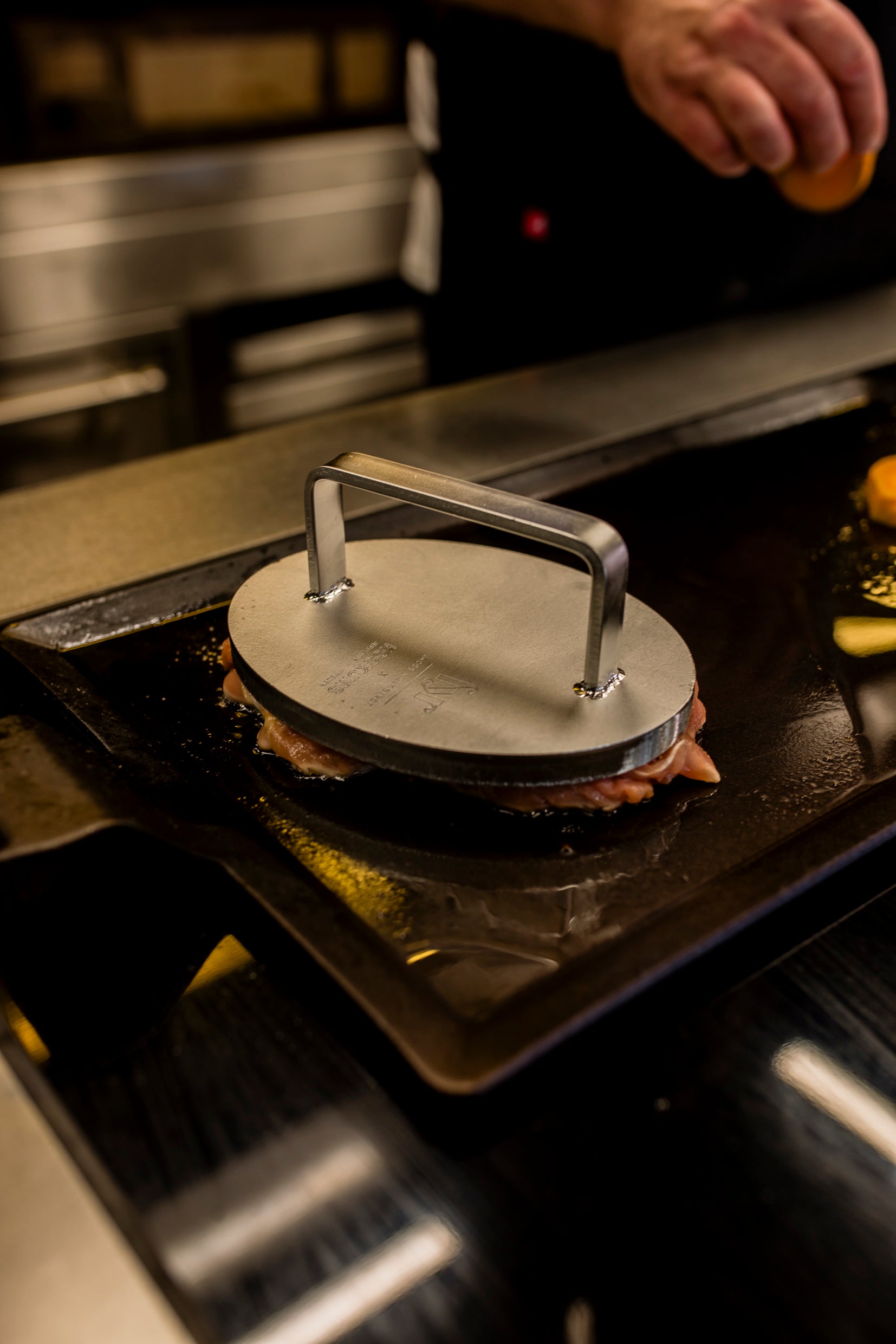 Das Kockums Jernverk EDELSTAHL-GRILLGEWICHT drückt auf das auf der Grillplatte brutzelnde Fleisch, eine Hand im Hintergrund sorgt für perfektes knuspriges Braten.