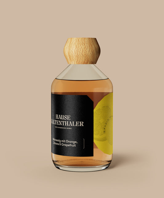 Eine Flasche ZITRUSESSIG mit Orange, Zitrone und Grapefruit aus dem Hause Kaltenthaler mit Holzverschluss und schwarzem Etikett mit Zitrusgrafiken steht elegant auf beigem Hintergrund; ideal zum Verfeinern Ihrer Lieblings-Salatdressings.