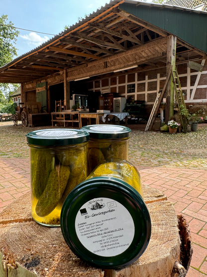 Drei Gläser EINGELEGTE GEWÜRZGURKEN werden auf einem Baumstumpf präsentiert, mit der charmanten Scheune des Biohofs Wilkenshoff in Niedersachsen als malerischer Kulisse.