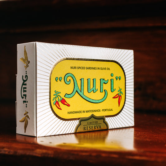 Eine Schachtel NURI PIKANTE JAHRGANGS-SARDINEN RESERVA 2020 in Olivenöl ruht auf einer Holzoberfläche und zeigt lebendige Grafiken und Texte, ein Beispiel für exquisite portugiesische Konservenproduktion.
