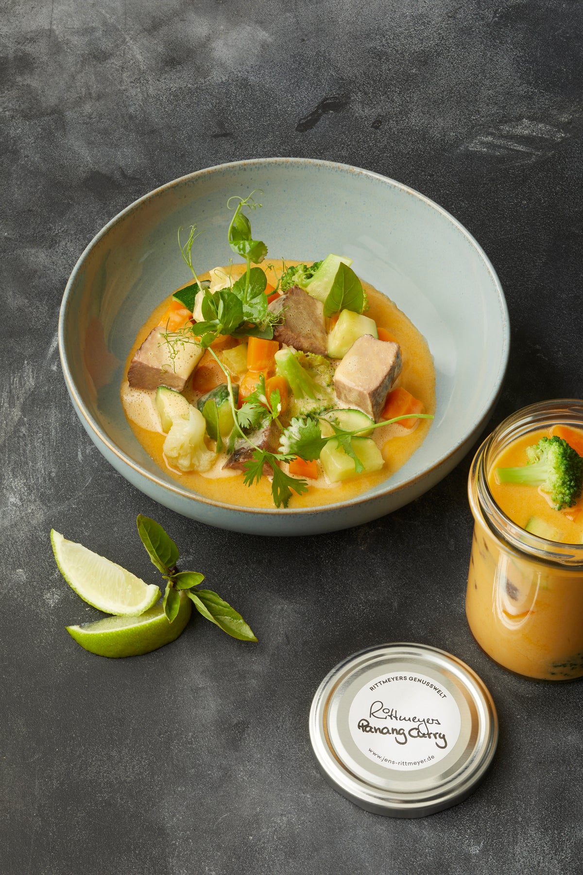 Eine Schüssel PANANG CURRY MIT LANGSAM GEGARTEM RIND mit zartem Fleisch und frischem Gemüse von Rittmeyers Besondere Raffinessen, begleitet von Limettenspalten und einem Glas dieser Köstlichkeit, steht einladend auf einer dunklen Oberfläche.