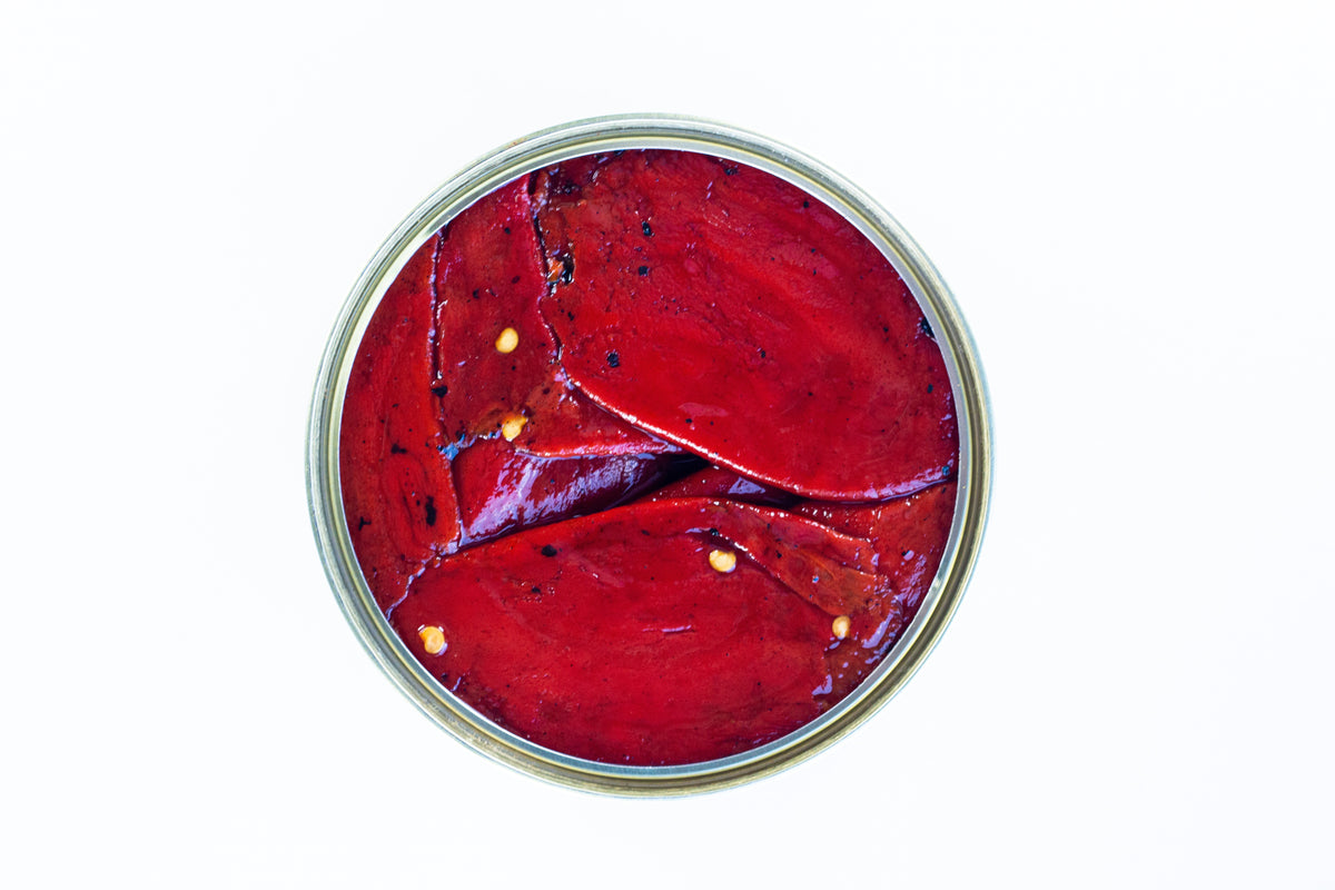 Draufsicht auf geschnittene EINGELEGTE PIQUILLO-PAPRIKA mit Samen, angeordnet in einem runden Glas vor weißem Hintergrund. Marke: El Carrascal.