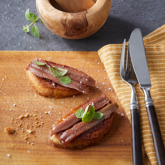 Zwei Bruschetta, belegt mit CODESAs handfiletierter Sardelle und duftendem Basilikum, liegen auf einem Holzbrett, neben Mörser und Stößel, Messer und Gabel.