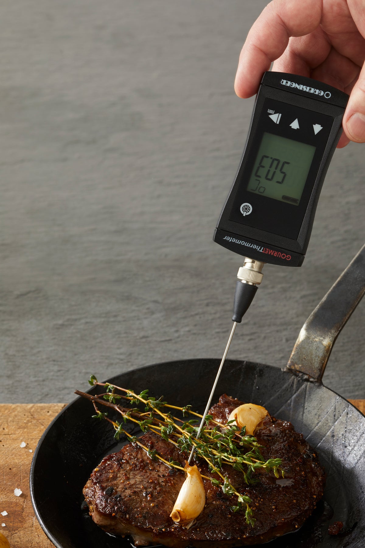 Ein DIGITALES GARTHERMOMETER von Greisinger misst die Temperatur eines Steaks in einer Pfanne, garniert mit Knoblauch und Kräutern.